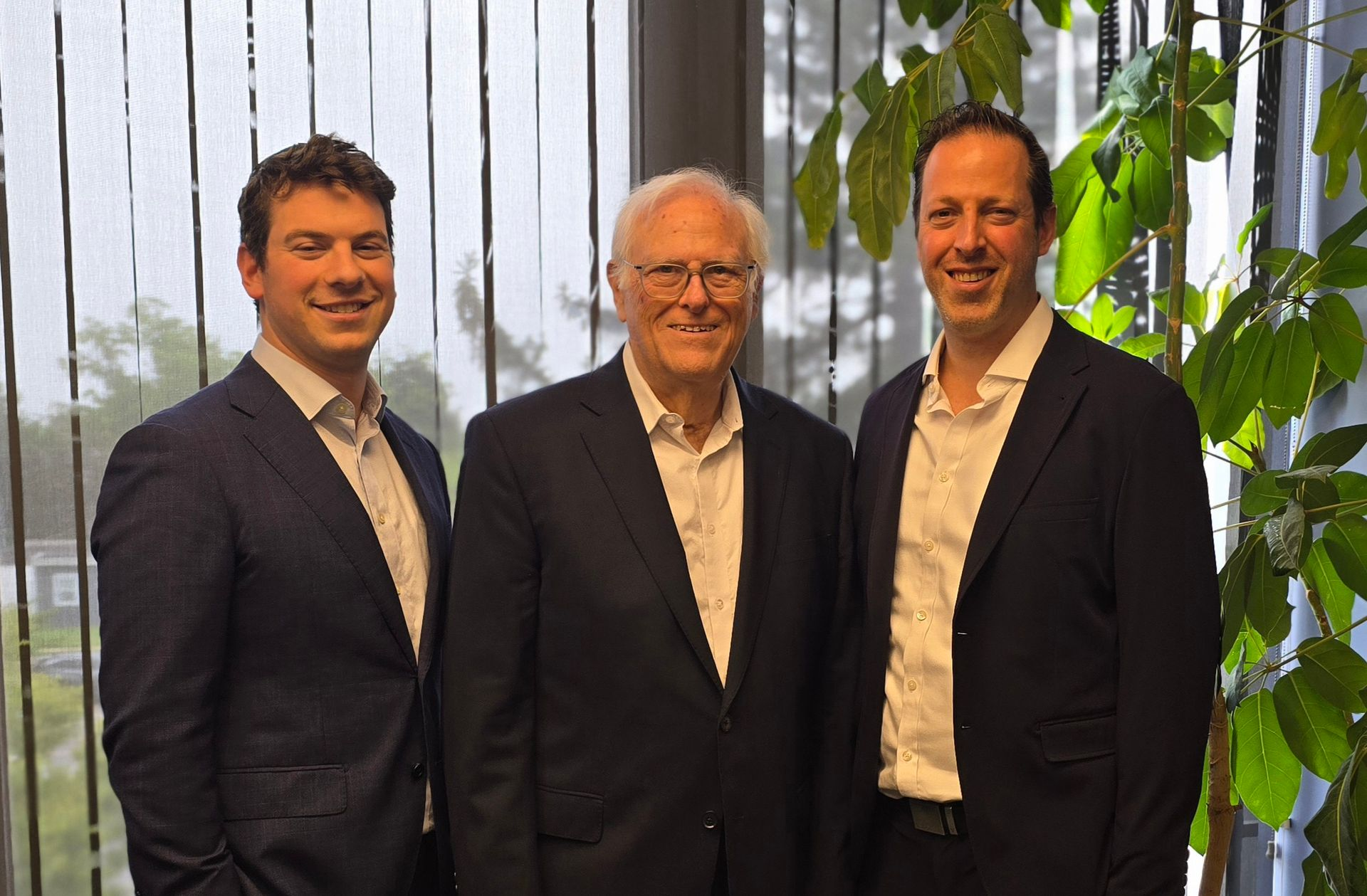 Three men in suits stand indoors near a window and plants; they are smiling at the camera.